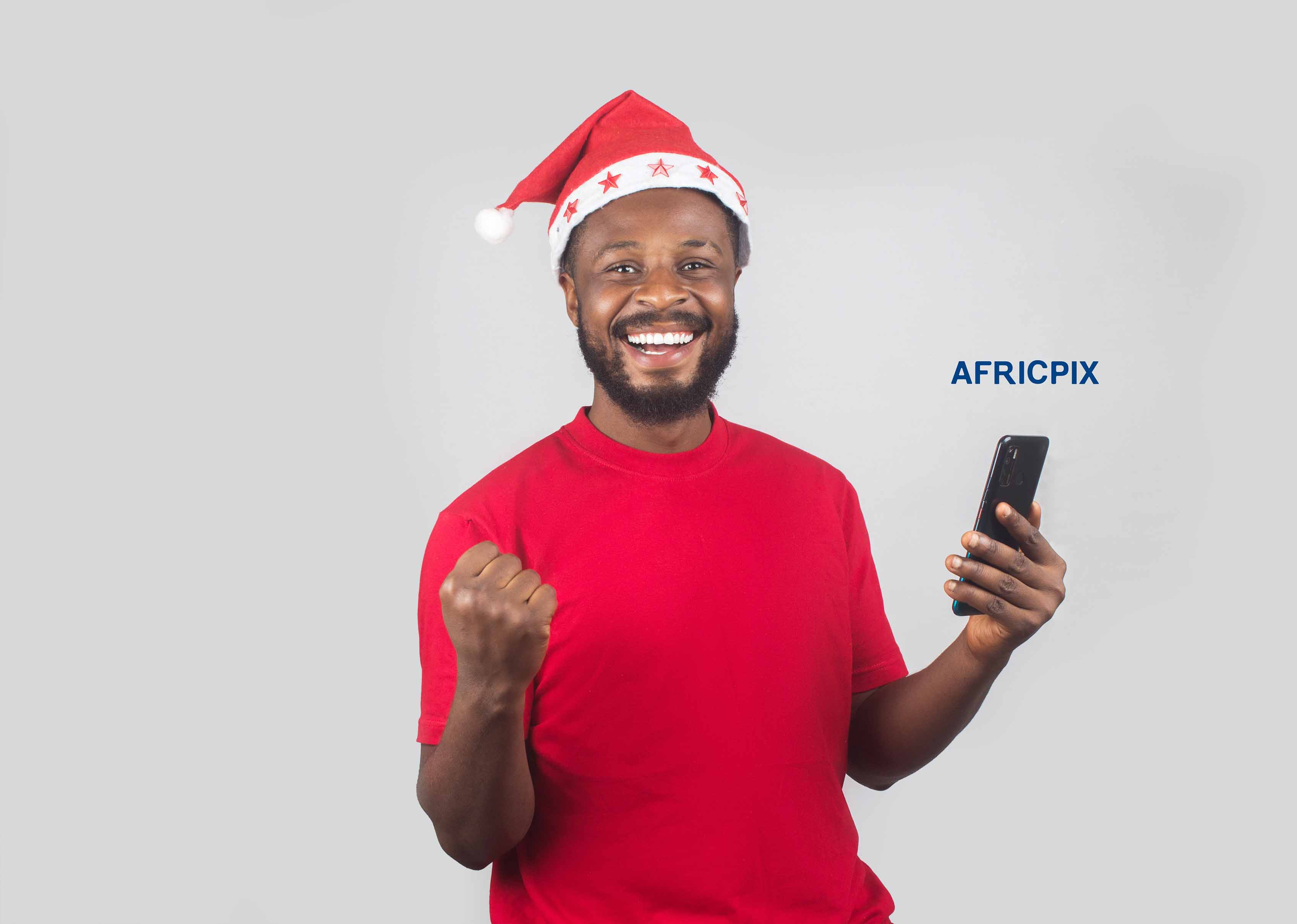 An African Nigerian man wearing a Christmas cap, holding a phone, and looking excited with a big smile.